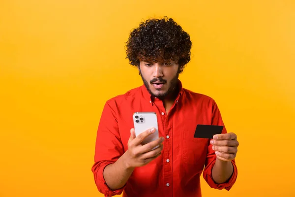 Потрясенный взволнованный мужчина с бородой, держащий кредитную карту и смартфон, смотрящий на экран смартфона большими глазами, удивленный легким банковским обслуживанием — стоковое фото