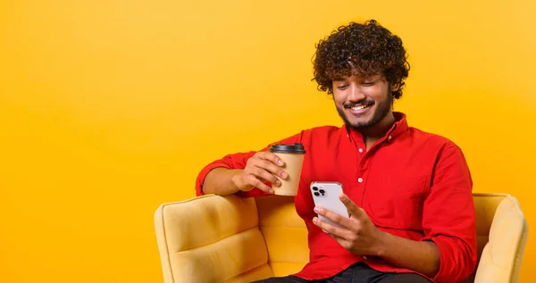 Portrait de blogueur mec indien isolé sur le mur orange tenant tasse de café, streaming vidéo en ligne via téléphone mobile, prendre selfie pour les réseaux sociaux, posant — Photo