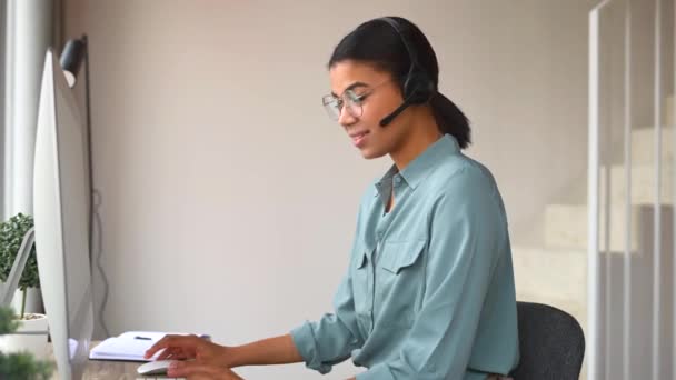 Jovem mulher alegre usando fone de ouvido para conversar on-line sentado na mesa no escritório em casa — Vídeo de Stock