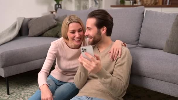 Couple in love are sitting on the floor at home and using smartphone — ストック動画