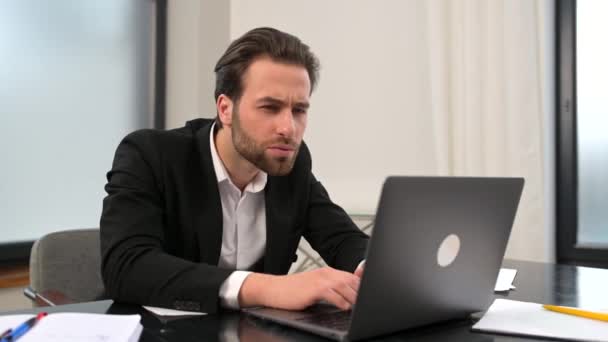 Zmatený mladý podnikatel sedí za stolem v kanceláři a dívá se na obrazovku notebooku — Stock video