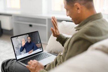 Hoş geldiniz. Gülümseyen ve neşeli genç adamın evdeki rahat koltukta bilgisayar kullanarak video araması yaparken yan görüntüsü. Günlük kıyafetli yakışıklı adam webcam 'e merhaba diyor.