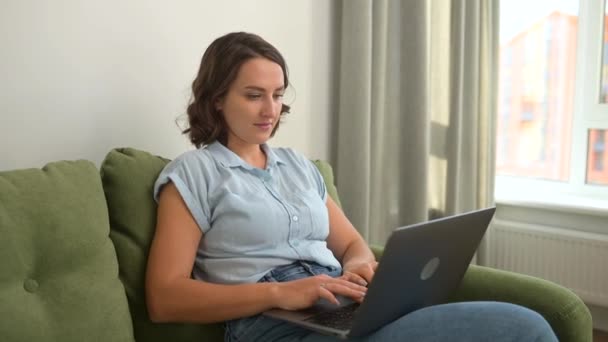 Atractiva mujer enfocada vistiendo camisa casual usando portátil sentado en casa en el sofá — Vídeos de Stock