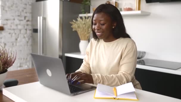 Una hermosa mujer afroamericana en ropa casual está usando una computadora portátil sentada en su oficina en casa — Vídeo de stock