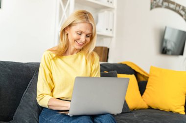 Modern olgun 60 'lı yaşlardaki kadınlar hafta sonlarını evde geçirmeyi, bilgisayar aygıtlarında kablosuz internet izlemeyi seviyorlar. Mutlu yaşlı kadın oturma odasında koltuğa otur, dizüstü bilgisayarla çalış.