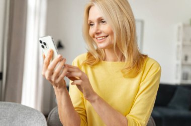 Glad mature woman talking online on mobile phone via video connection, participating in a conference, sitting at the table video-calling