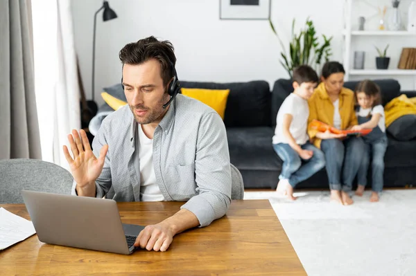 O pai ocupado está a trabalhar à distância. Homem usando laptop para trabalho freelance sentado à mesa em casa, enquanto a família sentado pacificamente — Fotografia de Stock