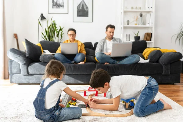 Father and mother working on the distance during quarantine. Parents work remotely with laptops sitting on the couch in living room, two sweet kids playing with toys — Stock Photo, Image