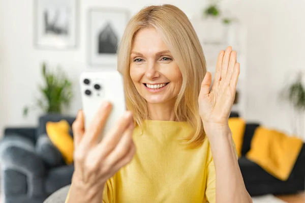 Feliz sorrindo mulher madura sentada na cozinha, relaxando e conversando com sua amiga ou família através de conexão de vídeo, fêmea sênior segurando telefone celular e acenando — Fotografia de Stock