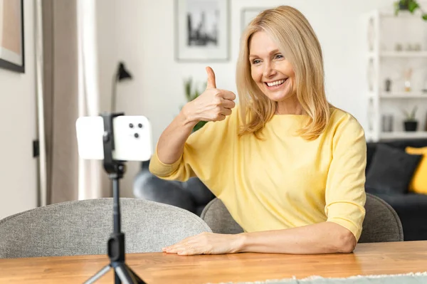 Senior attractive woman speaking to the camera on the phone on a tripod, gesturing, blogging or speaking live, recording a video presentation, holding video call —  Fotos de Stock
