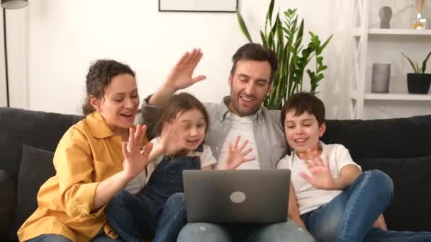 Virtual meeting. Family of four waving to screen while using laptop for video connection — стоковое видео
