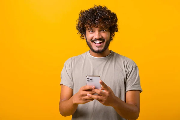 Экстатичный кудрявый молодой индиец, стоящий изолированный на желтом и использующий модный смартфон — стоковое фото