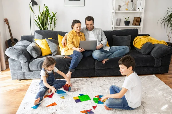 Ebeveynler karantina sırasında uzakta çalışıyor. Anne ve baba oturma odasındaki kanepede dizüstü bilgisayarlarla uzaktan çalışırlar. İki tatlı çocuk oyuncaklarla oynarlar. — Stok fotoğraf