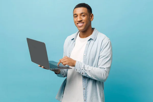 Optimistic african-american male student in casual jeans shirt using laptop pc isolated on blue — Stock fotografie
