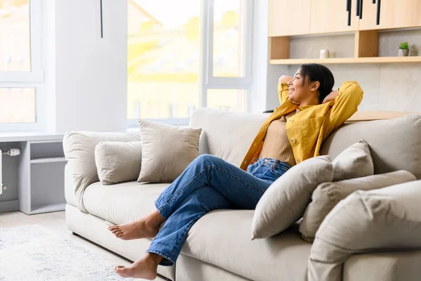 Charming and attractive young African-American woman resting, peaceful and serene multiracial lady looking at the camera sitting on the sofa in cozy living room, spends weekend — стоковое фото