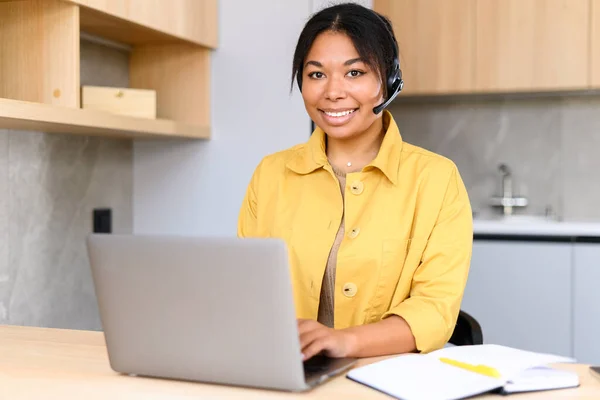 Saleswoman taking a call while working remotely from home office. Cheerful attractive woman wearing wireless headset using laptop for video connection — Foto de Stock