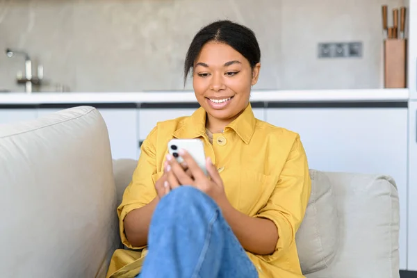 Szczęśliwa wielorasowa kobieta siedząca na kanapie i spędzająca czas online ze swoim smartfonem, SMS-em, przewijaniem wiadomości, udostępnianiem postów w sieciach społecznościowych — Zdjęcie stockowe