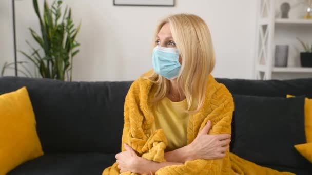 Konzept der Selbstisolierung. Aufgebrachte Frau mittleren Alters fühlt sich krank, sitzt in medizinischer Maske und mit Decke bedeckt — Stockvideo