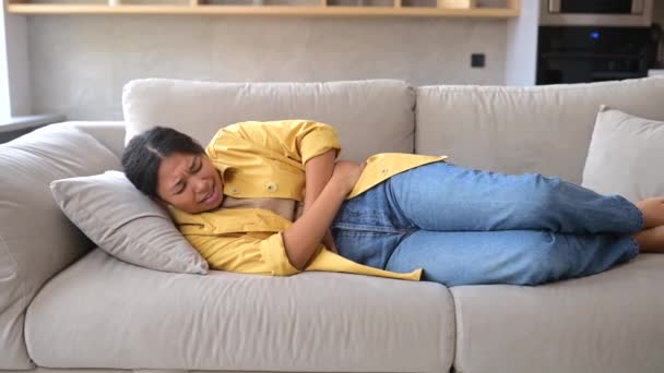 Young African-American woman sitting on the couch and suffers from stomach cramps — Stock videók