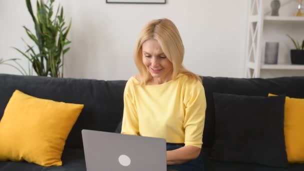 Iloinen vanhempi nainen istua sohvalla olohuoneessa, työskennellä kannettavan, hymyillen — kuvapankkivideo