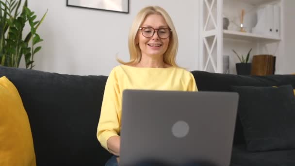 Confident 50s middle-age female teacher using laptop for virtual connection sitting on the sofa — Vídeo de Stock