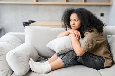 Üzgün kadın kanepede oturuyor, pencereden bakıyor, gelecek hakkında endişeli ve stresli hissediyor ve düşünüyor