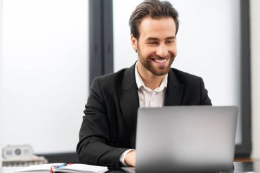 Resmi kıyafetler giyen neşeli beyaz adam masada oturuyor ve modern ofiste dizüstü bilgisayar kullanıyor.