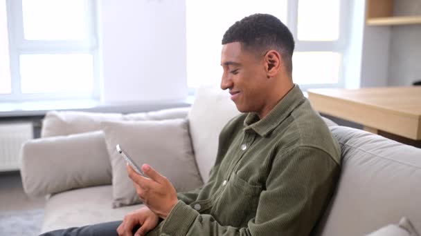 Alegre multirracial millennial chico sosteniendo smartphone sentado en el sofá en casa — Vídeos de Stock