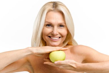 Happy senior woman holding fresh green apple middle her hands and smiling. Elderly model with naked shoulders posing and looking at the camera isolated on white