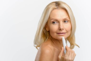 Yakışıklı sarışın kadın nemlendirici ruj kullanıyor, tedavi kremi kullanıyor. Mutlu ve iyimser orta yaşlı güzel kadın beyaza doğal dudak kremi sürüyor.