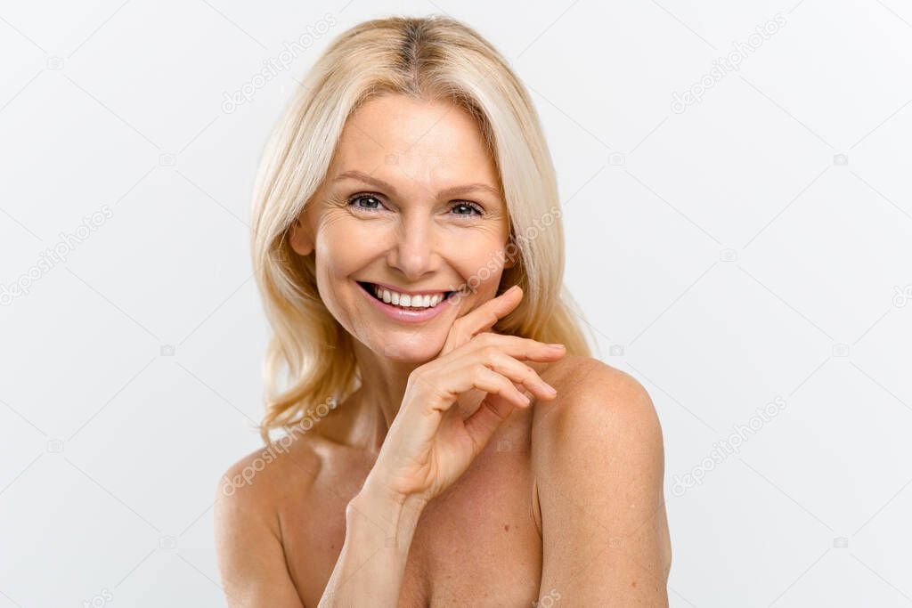 Portrait of fresh and gorgeous middle-aged woman with pure and healthy skin and blonde hair, stands with a naked shoulders isolated on white background, mature lady looks at camera