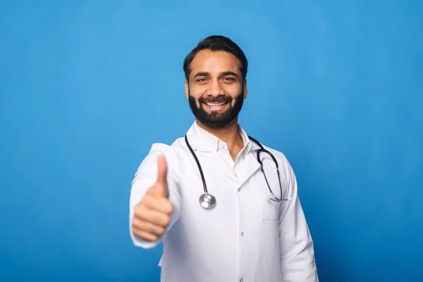 Cheerful Indian male doctor with a stethoscope in a medical gown posing isolated over blue — Stock Photo, Image
