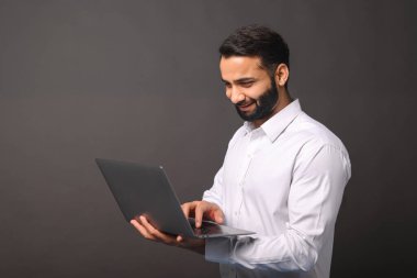 Resmi beyaz tişörtlü ve dizüstü bilgisayar kullanan mutlu ve sakallı bir Hintli adamın portresi.
