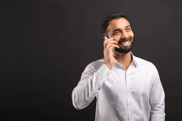 Happy bearded Indian man in formal white shirt talking on smartphone isolated on black — Stock Photo, Image