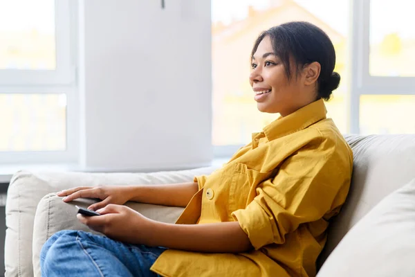 Boldog fiatal multiracionális nő tévéműsorokat néz, kényelmes kanapén ül távirányítóval, csatornát vált, nézi a képernyőt — Stock Fotó
