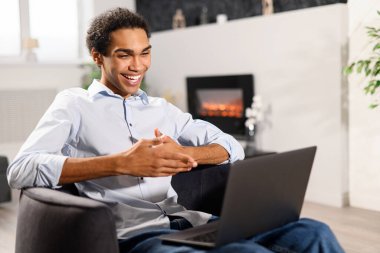 Video iletişimi, yakınlaştır. Çok ırklı genç adam evdeki bir video görüşmesi için dizüstü bilgisayar kullanıyor, gülümseyerek webcam 'e bakıyor.