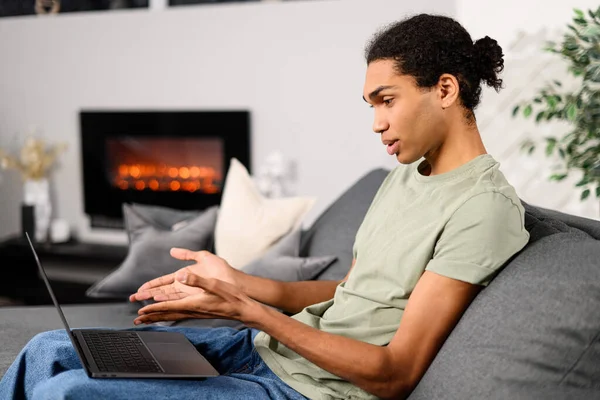 Sanal toplantı, video çağrısı konsepti. Rahat koltukta oturan neşeli, çok kültürlü bir adam interinterinterlocutor ile konuşurken video görüşmesi için son moda dizüstü bilgisayarını kullanıyor.