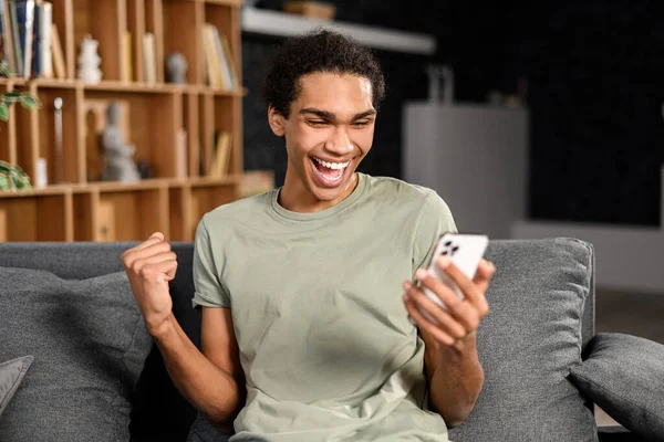 Ενθουσιασμένος πολυφυλετικός άντρας κάθεται στον καναπέ κοιτάζοντας το τηλέφωνο και χαμογελώντας, έκπληκτος συναισθηματικός τυχερός άνθρωπος, χαίρεται με το καλό μήνυμα, ειδήσεις — Φωτογραφία Αρχείου