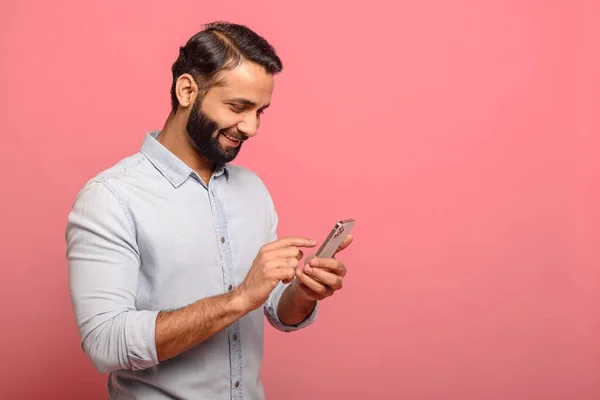 Happy Indian man in casual jeans shirt using smartphone isolated on pink — стоковое фото