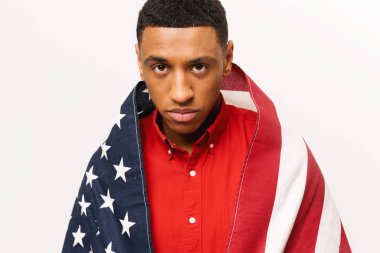 Serious worried African-American guy stands covered with american flag isolated over white background