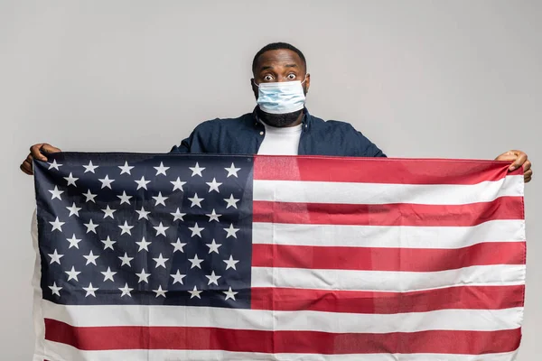 Surprised African-American man in protective medical mask holding USA flag — стокове фото