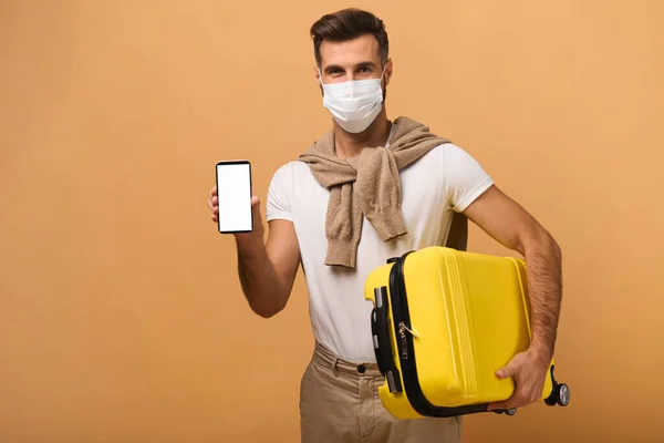 Ευτυχισμένος άνθρωπος φορώντας προστατευτική μάσκα κρατώντας smartphone με άδεια οθόνη και δείχνοντάς το στην κάμερα ενώ ποζάρει με τις αποσκευές του απομονωμένες σε πορτοκαλί — Φωτογραφία Αρχείου