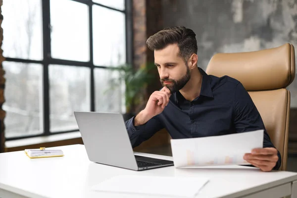 Concentrated male small business owner holding papers and looking at the laptop screen while sitting in modern loft office. Successful man manager in casual wear looking at the document — Stok fotoğraf