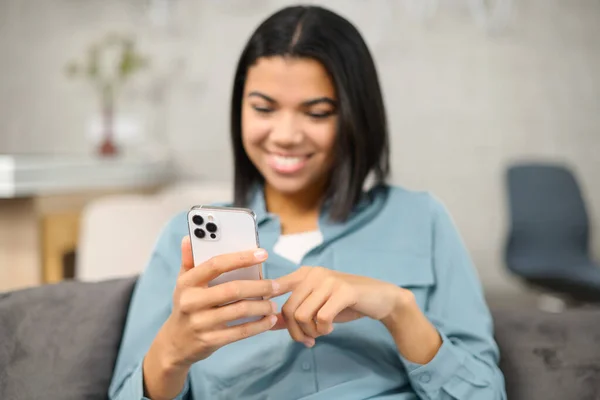 Cheerful young woman using cell phone for chatting via messages while sitting down on the comfortable couch at home. Smiling student chatting with classmates online — стоковое фото