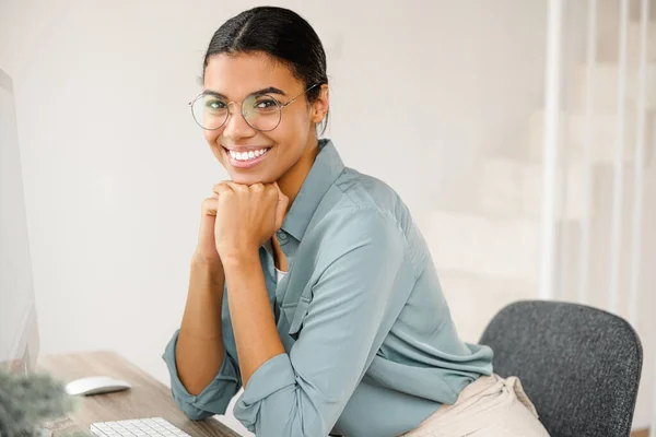 Pozitivní inteligentní mladá smíšená-závod studentka se usmívá, zatímco sedí u stolu s počítačem, studuje z domova, sledování on-line webinář — Stock fotografie