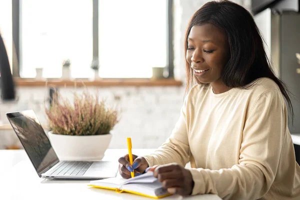 Uma mulher afro-americana bonita no desgaste casual está usando o computador portátil que se senta no escritório home — Fotografia de Stock
