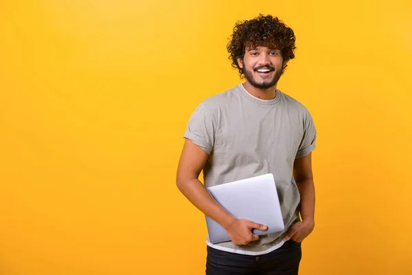 Cheerful Indian young man holding laptop standing isolated on yellow — Stock fotografie