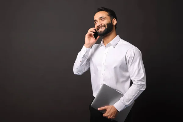 Busy handsome confident bearded Indian man in formal wear talking on smartphone carrying laptop — Stock Photo, Image