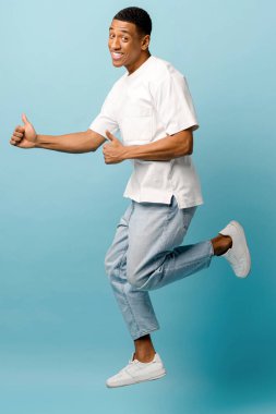 Heyecanlı Afro-Amerikalı genç adam mavi arka planda zıplıyor, stüdyoda eğleniyor.