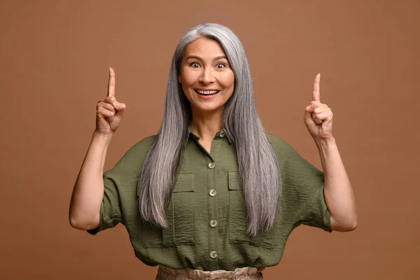 Smiling mature grey-haired female business woman isolated on grey background points finger up. Lady paying attention — ストック写真